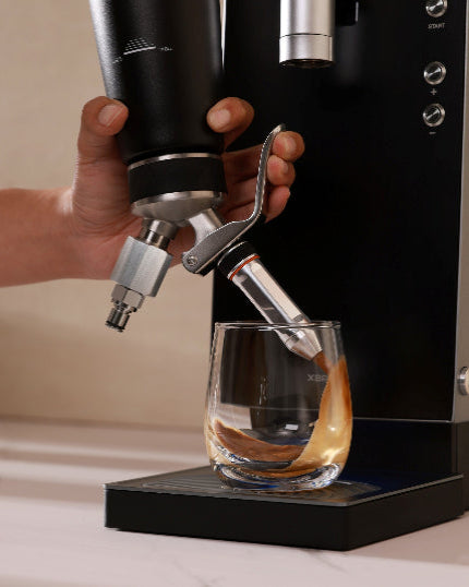 Nitro cream dispenser being used to pour coffee into a glass on a light-colored surface.